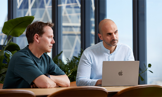 Ahmed og en kollega sitter ved et bord med MacBooken foran seg. Han ser på noen utenfor bildet.