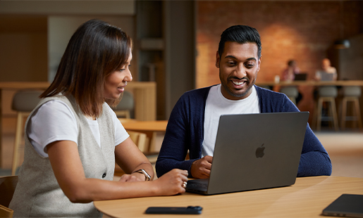 Amar sitter ved et bord i fellesområdet og samarbeider med en kollega på MacBooken sin.