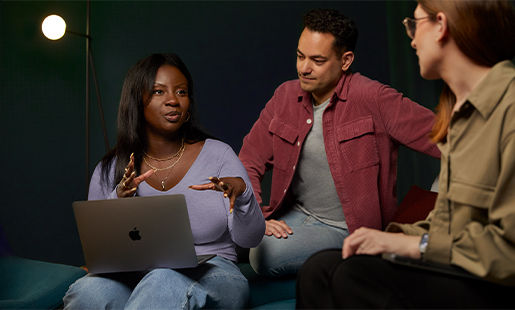 Ginelle sitter med en MacBook på fanget og samarbeider med to kollegaer.