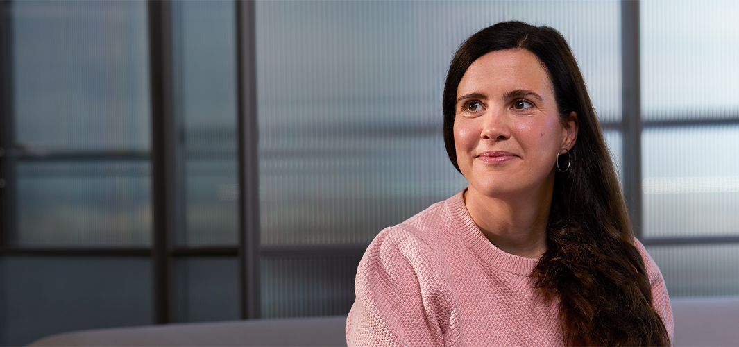 Lucie sitter smilende innendørs foran en delvis gjennomsiktig glassvegg og ser mot høyre.