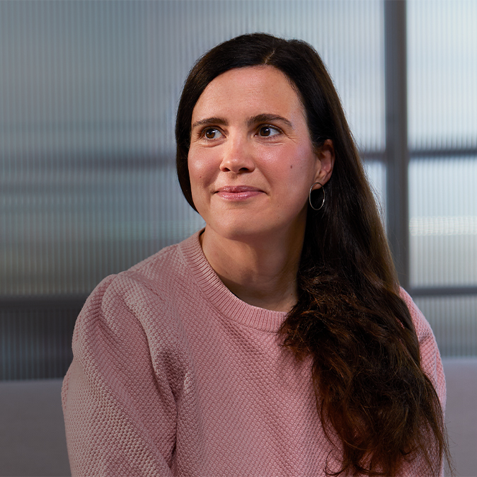Lucie sitter smilende innendørs foran en delvis gjennomsiktig glassvegg og ser mot høyre.