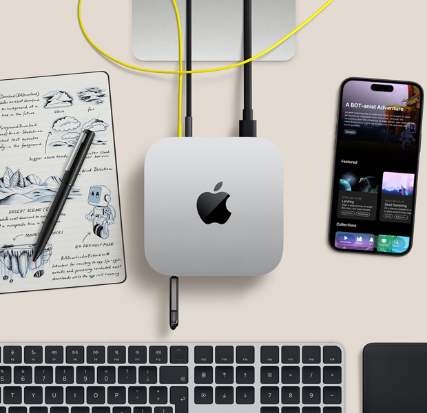 Vista desde arriba, Mac mini, aluminio color plata, esquinas redondeadas, logo de Apple negro brillante en el centro, conectada con un cable, memoria USB externa en el puerto USB-C frontal, base de un monitor Studio Display parcialmente visible, iPhone, Magic Trackpad, Magic Keyboard con Touch ID, pluma y bloc de notas de tinta electrónica
