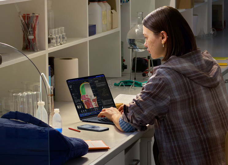 Una persona sentada en un laboratorio usando un MacBook, cuya pantalla muestra varias apps.