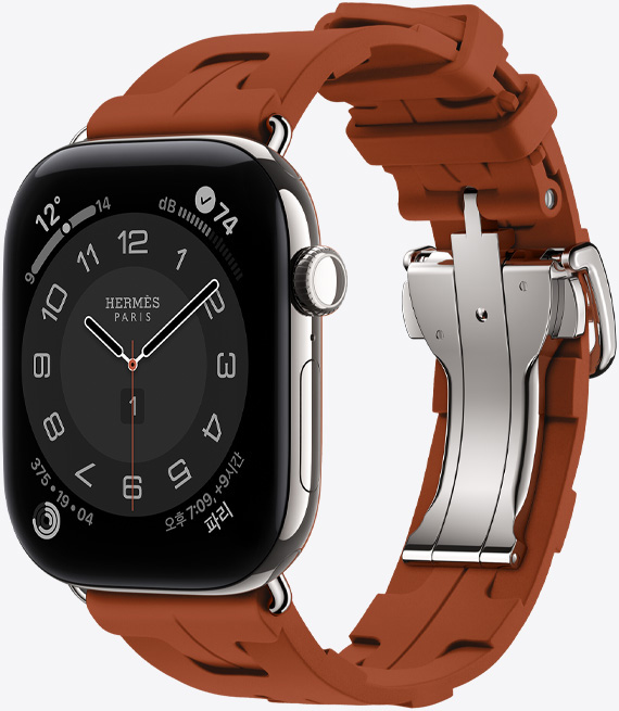 Apple Watch Hermès Series 12, Circulaire 아날로그 시계 페이스 및 시, 분, 초침, 컴플리케이션에 날씨, 소음, 활동 링, 파리 시간이 표시되어 있는 모습. 실버 색상 티타늄 케이스의 오른쪽 옆면 외관에 Digital Crown과 측면 버튼을 보여주는 모습. 뀌브르(브라운) 색상 Kilim 싱글 투어 밴드와 연결되어 있고, 성형 고무 소재로 제작된 기하학적으로 맞물리는 디자인에 실버 색상 금속 클라스프가 달려 있는 모습