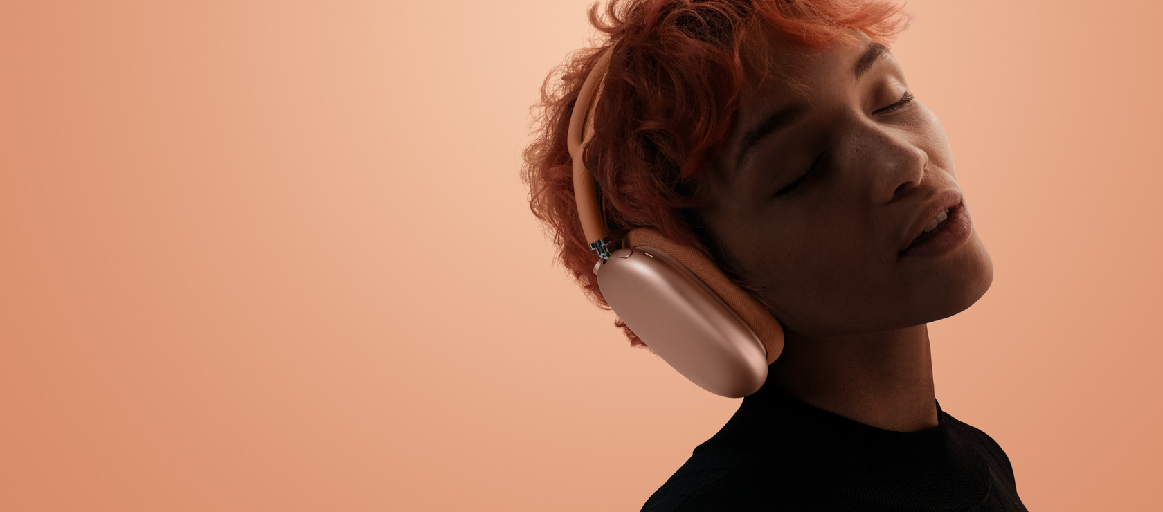 Osoba zatvorenih očiju i nagnute glave nosi narančaste slušalice AirPods Max 2, na narančasto-ružičastoj pozadini