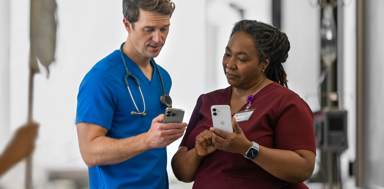 En mandlig sundhedsmedarbejder i blå uniform kigger på sin iPhone og viser noget på den til en kvindelig sundhedsmedarbejder i rød uniform, som også har en iPhone.